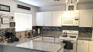 Una cocina moderna con armarios blancos, encimeras de granito, estufa a gas y microondas. Hay taburetes de bar y una ventana con persianas.