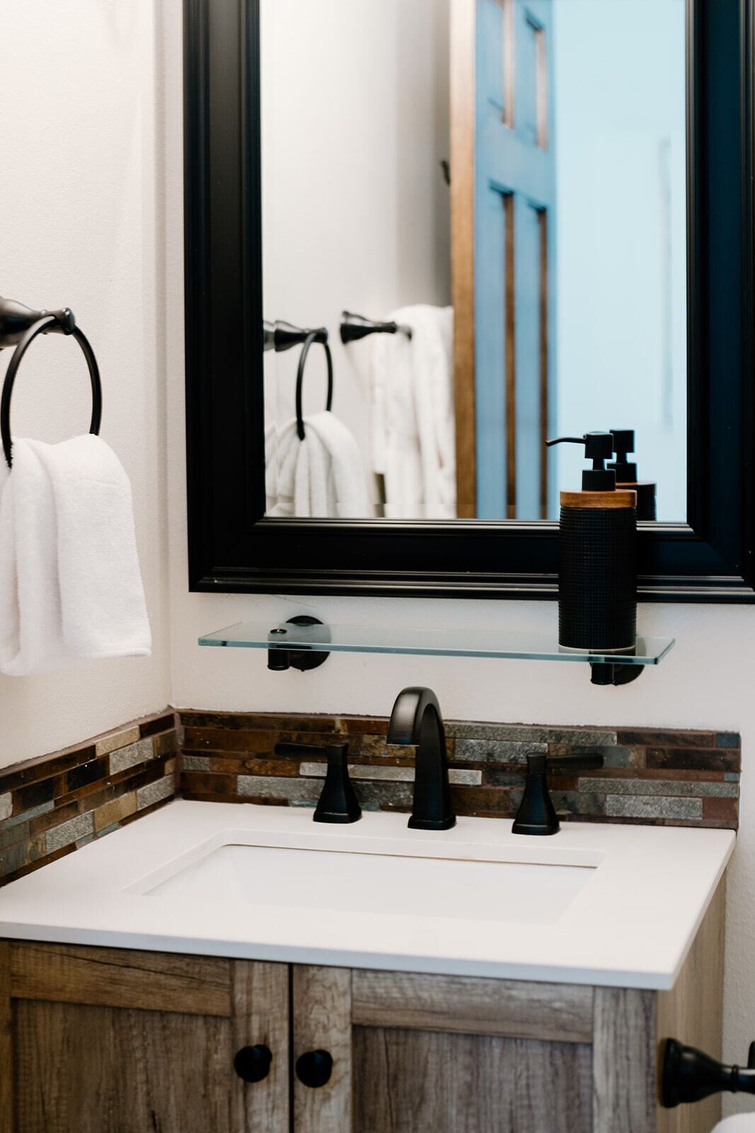 A bathroom with a white sink, black faucets, a glass shelf with a soap dispenser, and a mirror with a blue door reflection.
