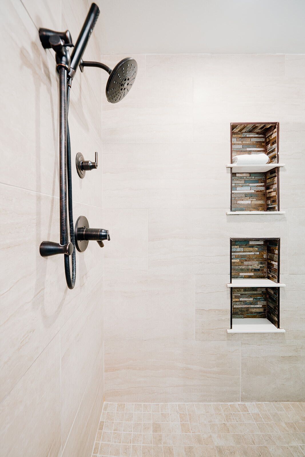Una ducha moderna con cabezal de ducha manual y un cabezal de lluvia, montados en una pared con baldosas de tonos claros. Hay dos repisas con diseño rústico, posiblemente para toallas o artículos de tocador.