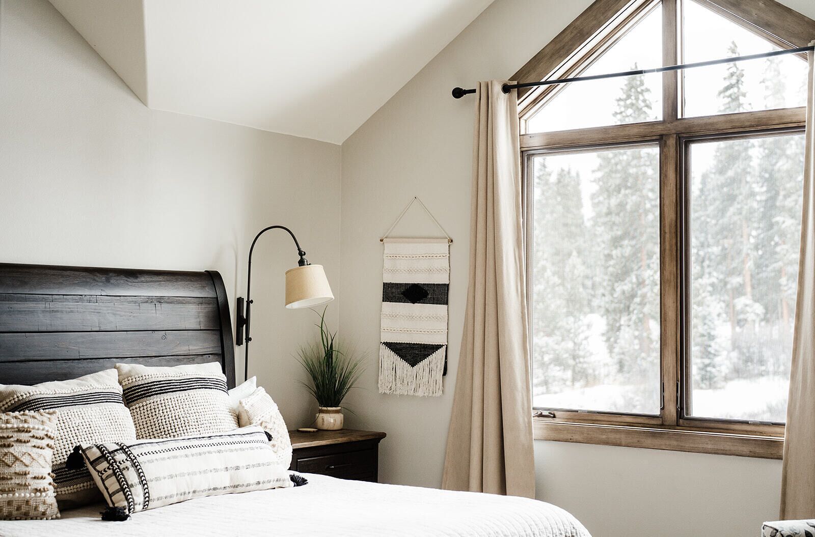 Ein Schlafzimmer mit Holz-Kopfteil, einer Nachttischlampe, einer Blumentopf-Pflanze, einer Wanddekoration und einem großen Fenster mit Blick auf schneebedeckte Bäume.