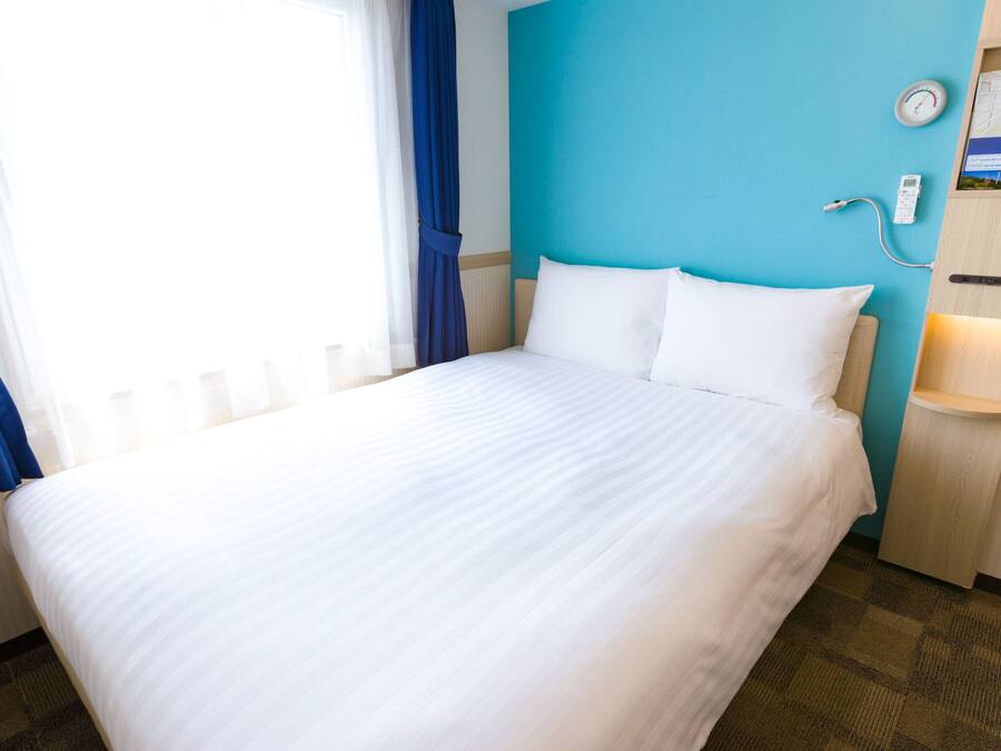 A hotel room with a bed, a nightstand, a clock, and a thermostat.