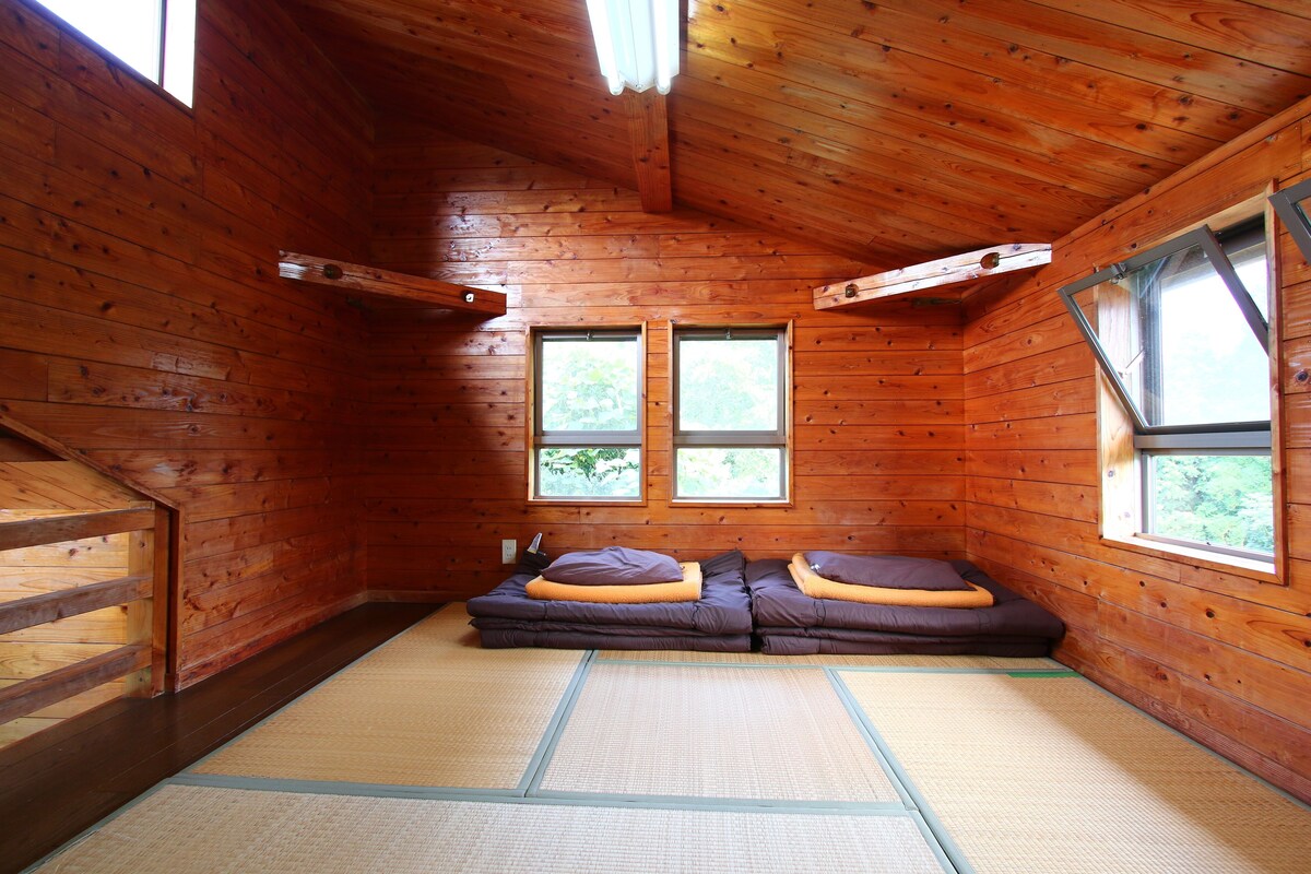 Una habitación de cabaña de madera con suelo de esteras tatami, dos ventanas y un sofá con cojines.