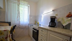 Una pequeña cocina con lavamanos, estufa y cafetera. Se observa una ventana con cortinas y una mesa con mantel estampado.