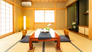 Ein traditioneller japanischer Raum mit Tatami-Boden, einem niedrigen Holztisch mit Unterlage, zwei Holzstühlen und einer Schiegetür mit dekorativem Paravan.