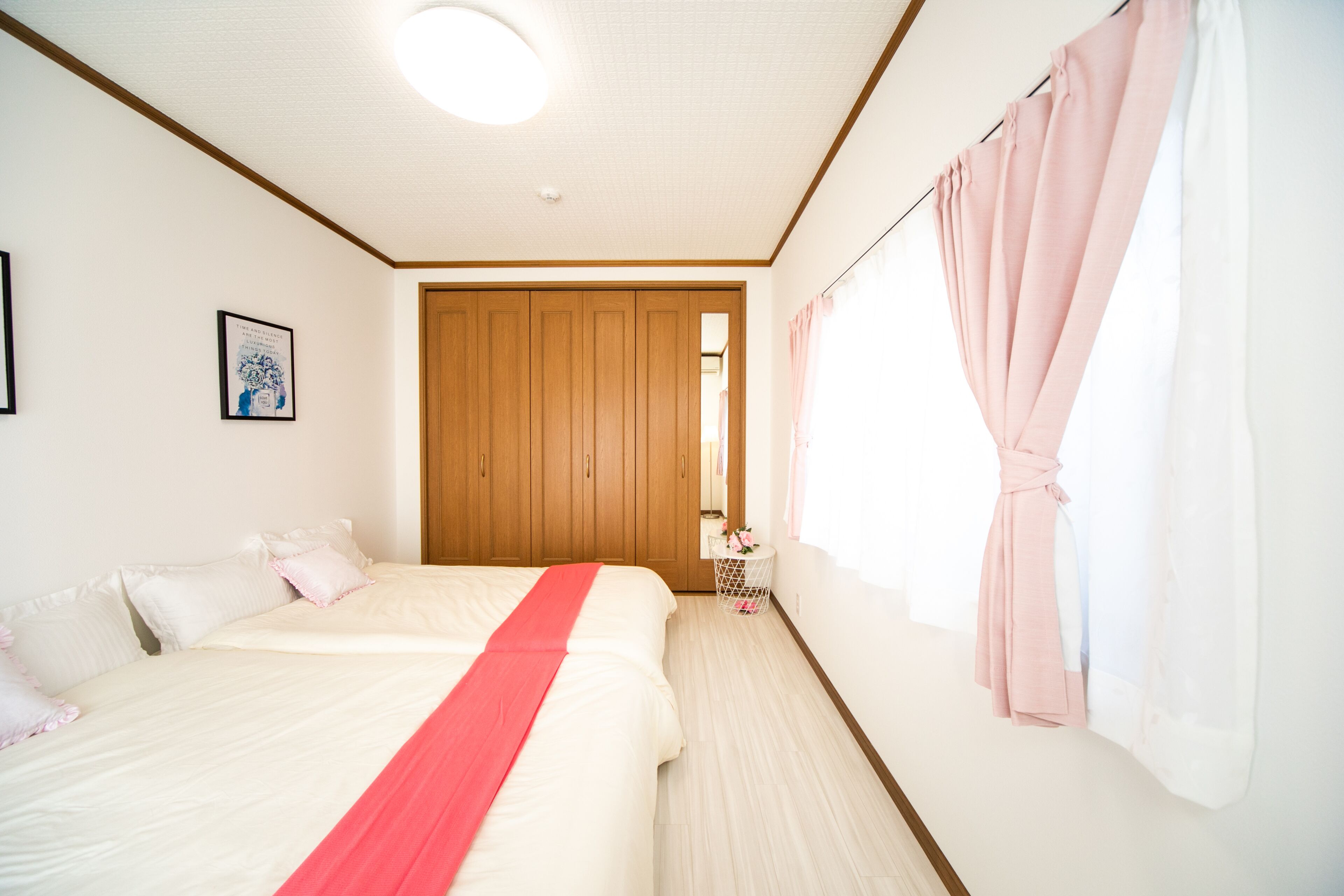 A bedroom with two beds, a wooden wardrobe, a small round table with a pink towel, and a window with pink curtains.