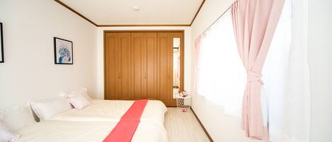A bedroom with two beds, a wooden wardrobe, a small round table with a pink towel, and a window with pink curtains.