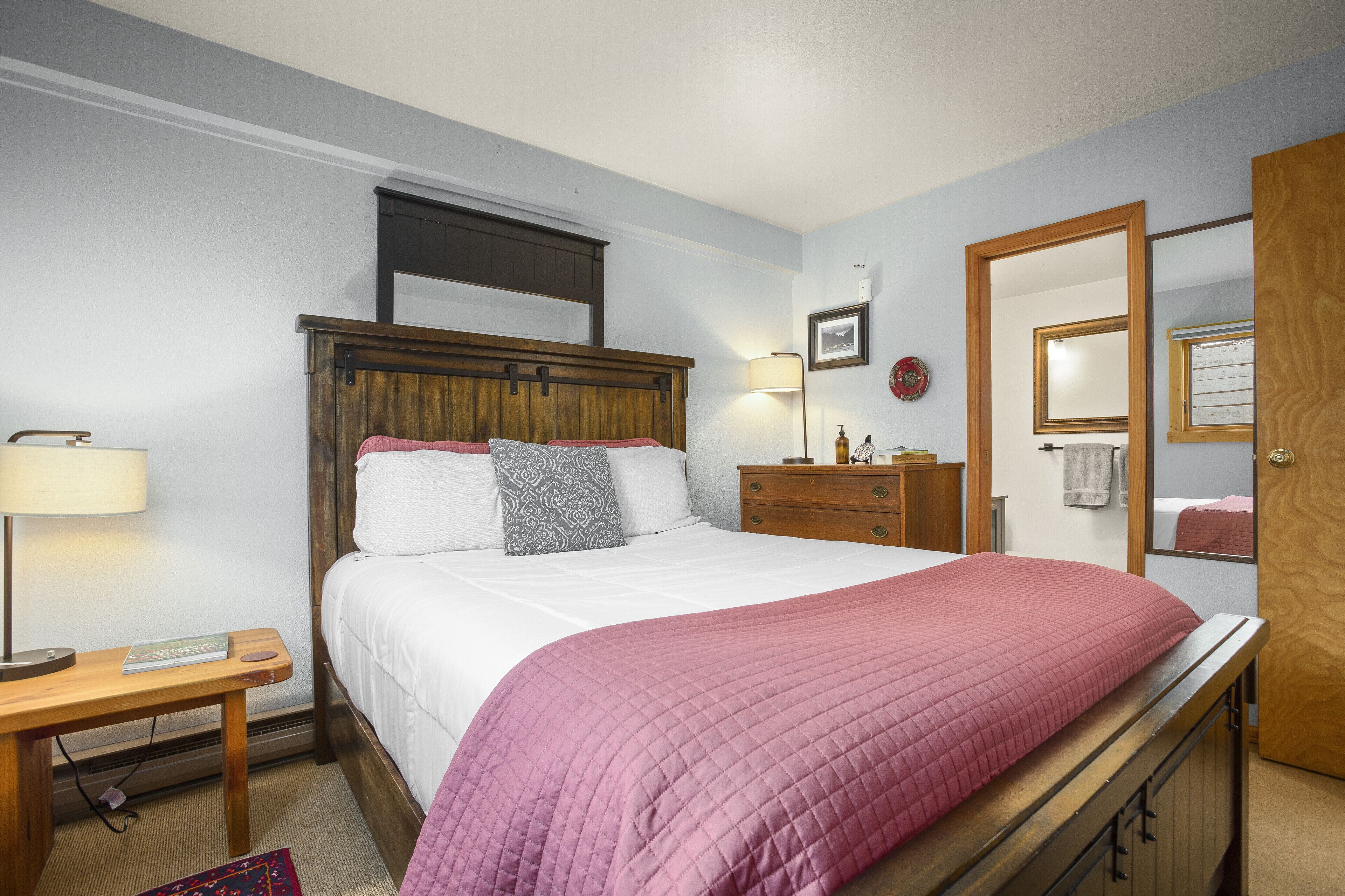 A bedroom with a bed, bedside table, lamp, dresser, mirror, and a door.