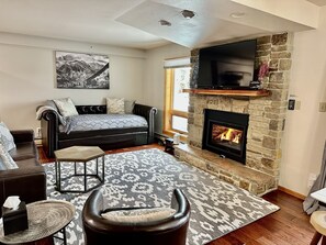 A cozy living room with a stone fireplace, a flat-screen TV mounted above it, a comfortable sofa, and a patterned rug. - Charming Lower Unit Condo: Stays Cool in Summer! (Steamboat Springs)