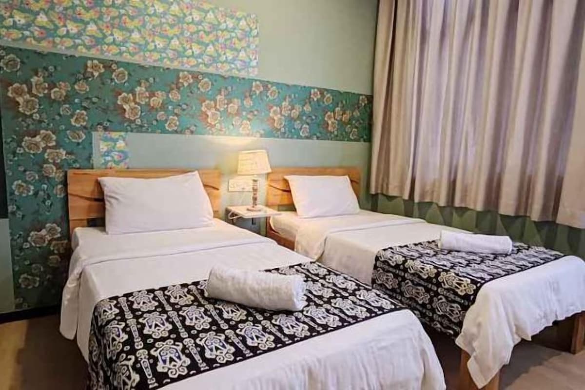 A hotel room with two beds, a nightstand with a lamp, and a floral-patterned wall.
