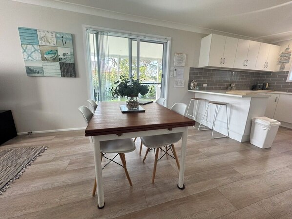 A modern kitchen with white cabinets, a wooden dining table, and a large window. - Pearly Shells Beach Cottage- 200m walk to Beach & 500m to shops - pet friendly (Culburra Beach)