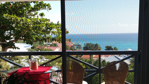 Un balcon avec vue sur l’océan, des fauteuils en osier, une table en verre et une nappe rouge.