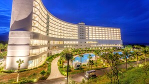 A modern hotel with a curved facade, multiple floors, and a central swimming pool surrounded by palm trees and greenery. - Selectum Noa Resort Cam Ranh (Cam Lam)
