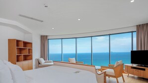 A modern hotel room with a large bed, a TV, a wooden cabinet, and a seating area with a view of the sea.