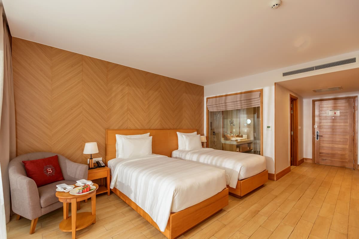 Une chambre d’hôtel avec deux lits, un canapé, une petite table avec une lampe et une salle de bain visible à travers une porte en verre.