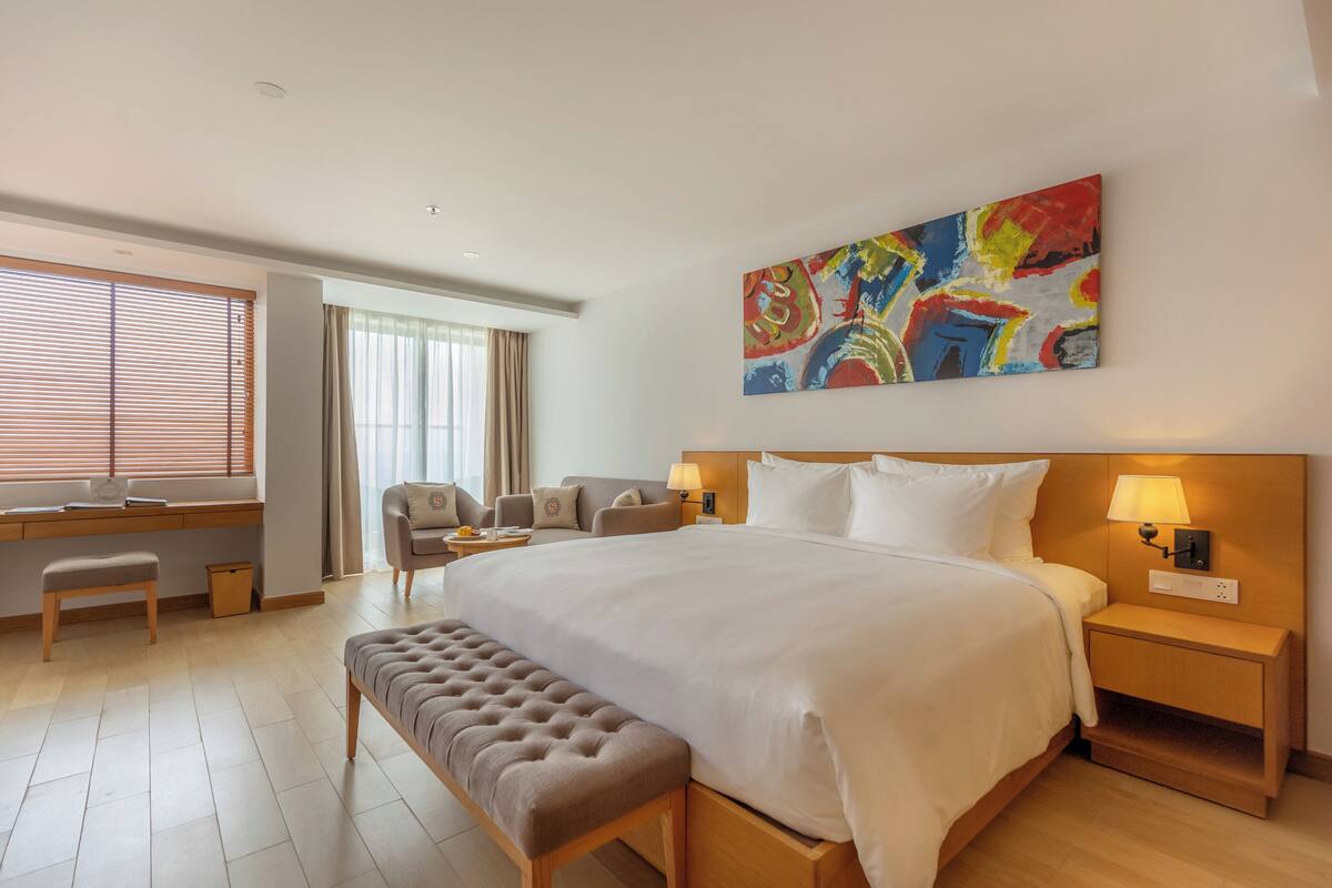 Une chambre d’hôtel avec un grand lit, un bureau avec une chaise, un banc, une table de chevet avec une lampe et une peinture abstraite aux couleurs vives accrochée au mur.