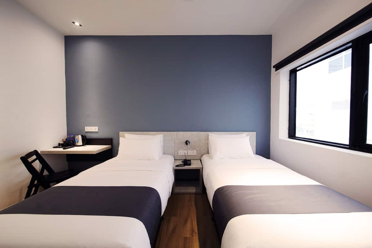 Une chambre d’hôtel avec deux lits simples, un petit bureau avec une chaise, une table de chevet et une fenêtre donnant sur la ville.