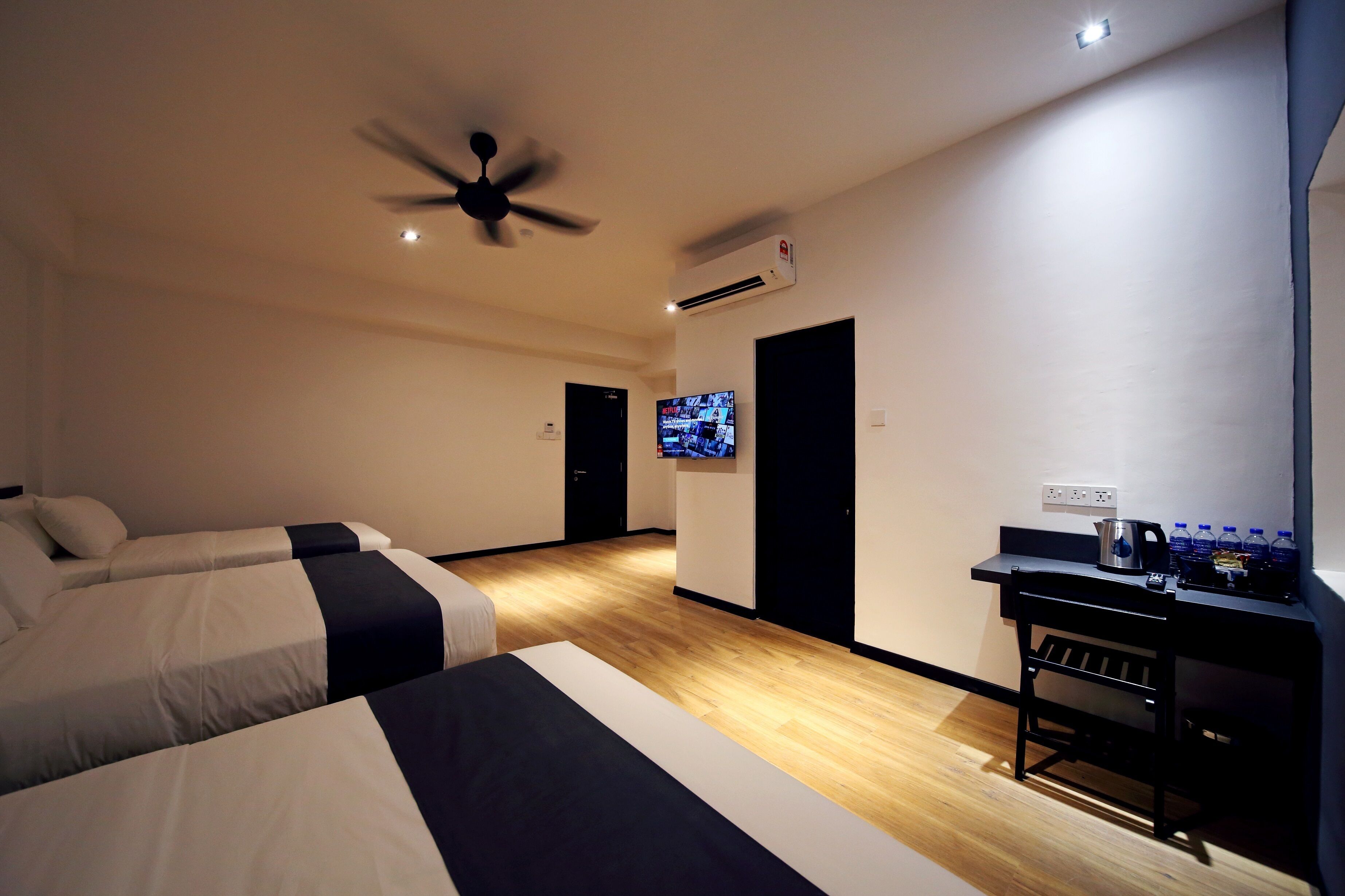 A hotel room with two beds, a desk with a kettle, a flat-screen TV, and a ceiling fan.
