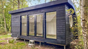 Une cabane moderne dotée de grandes fenêtres, d’une façade sombre et située dans un cadre forestier naturel.