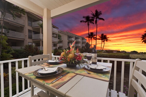 Une table à manger dressée pour deux personnes, avec vue sur le coucher de soleil, des chaises blanches et un arrangement floral tropical en guise de centre de table.