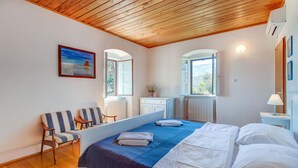 Um quarto com teto de madeira, uma cama com roupa de cama branca e azul, uma cadeira, uma mesa de trabalho e uma janela com vista para árvores.