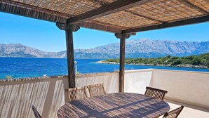 An outdoor patio with a round wooden table and chairs, a thatched roof, and a view of the sea and mountains.