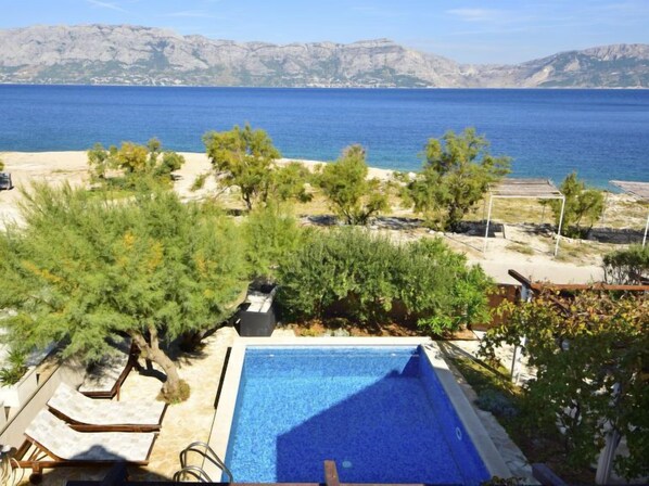 Un espace piscine agrémenté de chaises longues, entouré d’arbres et offrant une vue sur la mer et les montagnes.