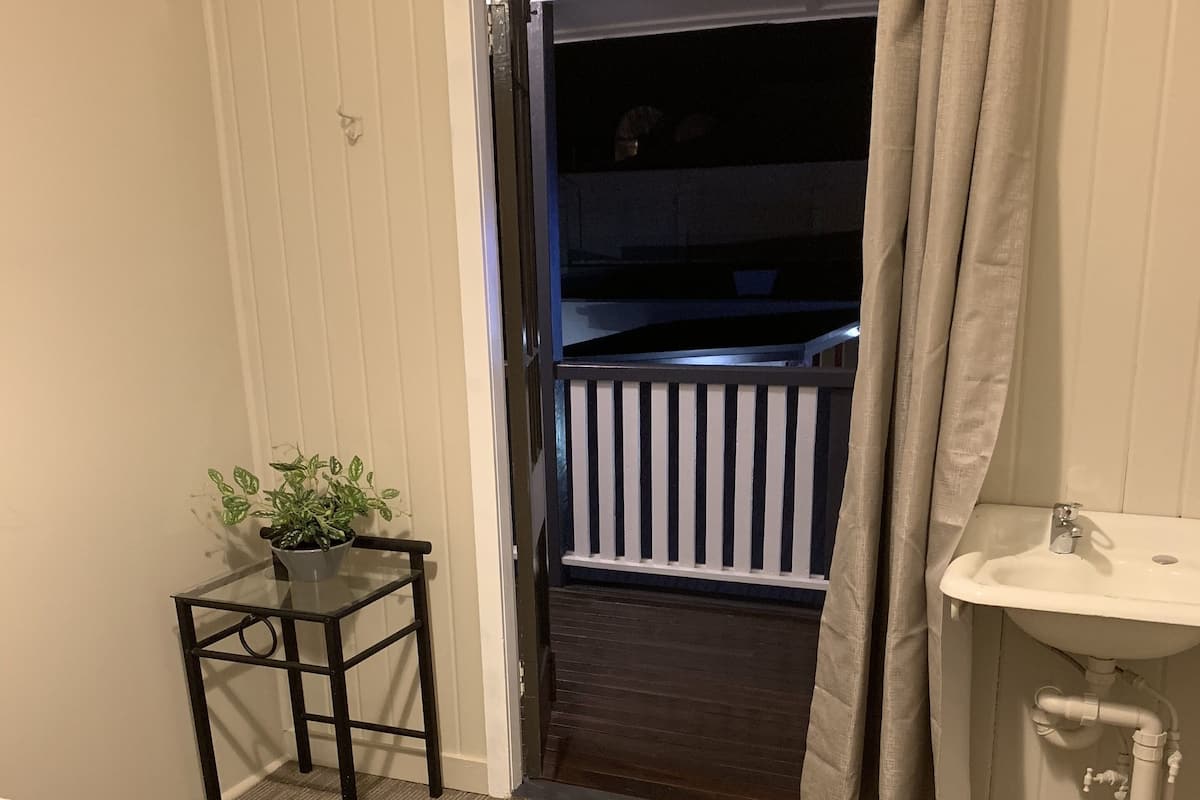 A hotel room with a bed, a small table with a plant, a bathroom with a sink, and a balcony with a railing.