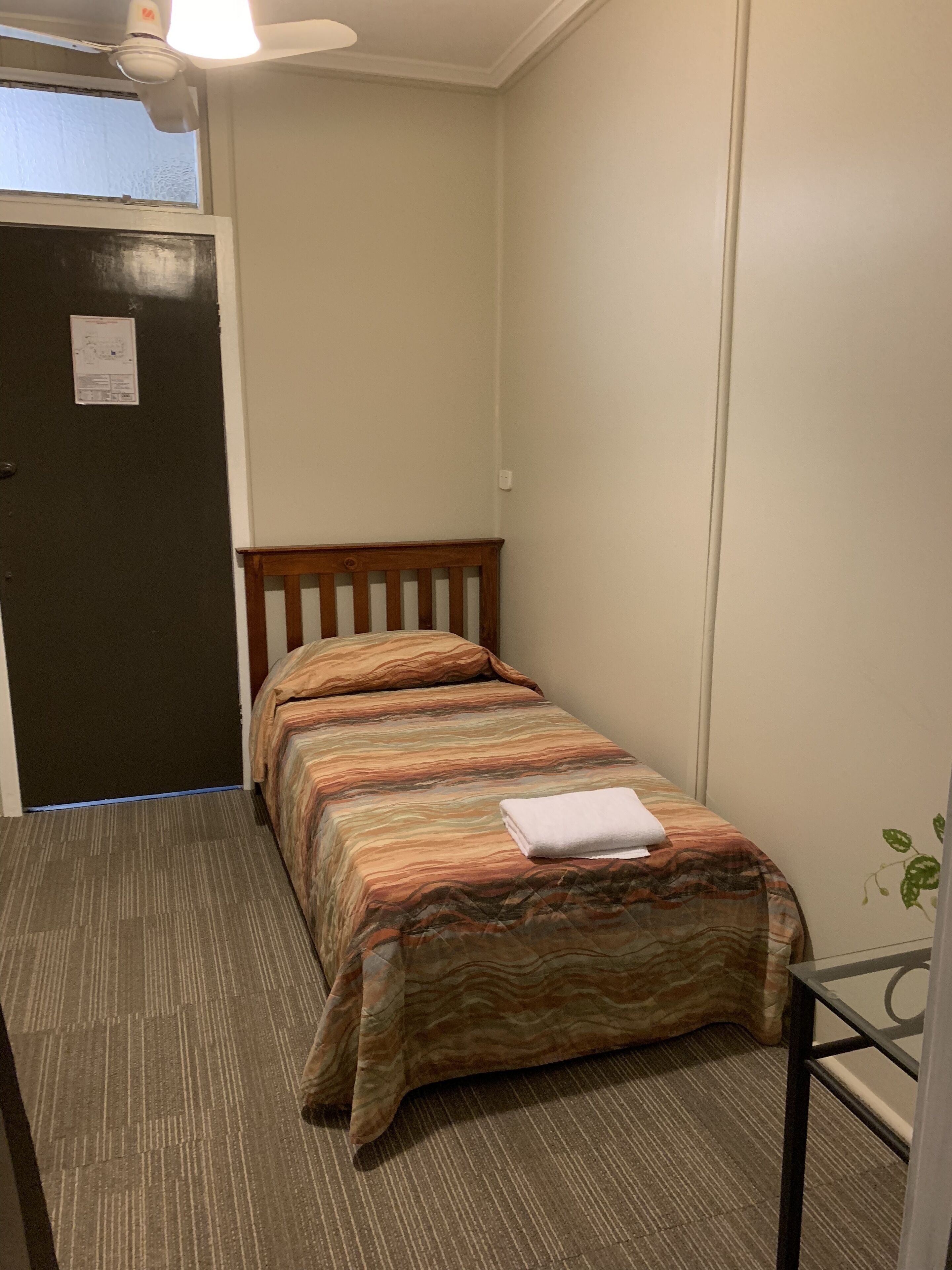 Una habitación pequeña con una cama individual, cabecera de madera, colcha estampada y una toalla doblada sobre la cama.