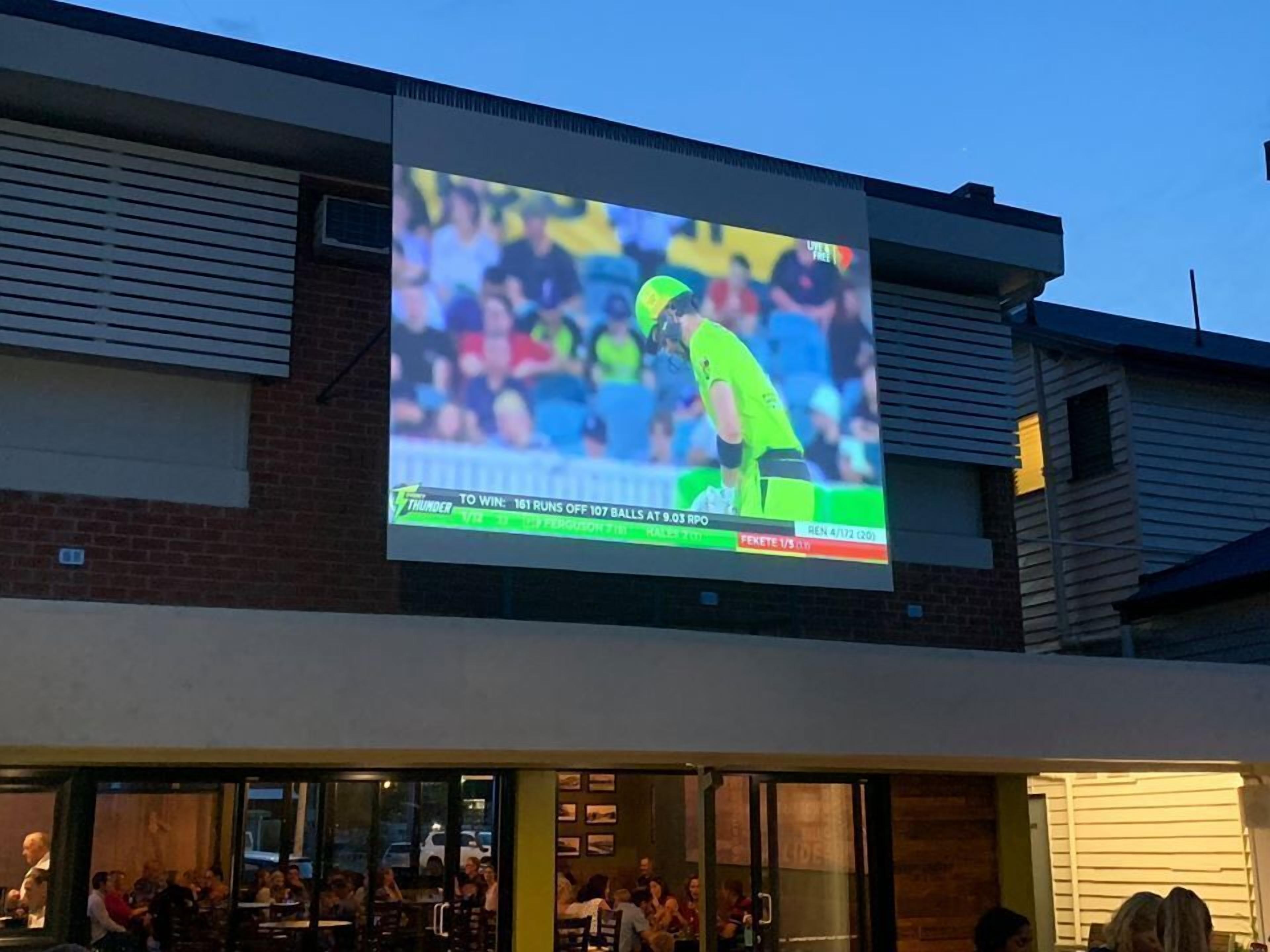 A sports event on a large screen at a venue with a brick facade and a flat roof.