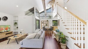 Ein geräumiges Wohnzimmer mit grauem Sofa, Couchtisch aus Holz und einem Fernseher. Eine Treppe mit Holzwandungen führt auf eine obere Ebene.