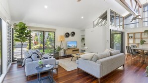 Ein geräumiges Wohnzimmer mit grauem Sofa, einem Couchtisch und einem Fernseher. Ein Balkon mit Pflanzen und Blick ins Freie ist ebenfalls vorhanden.