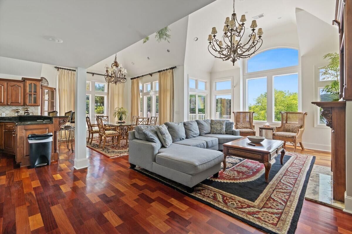 A spacious living room with a large sectional sofa, a dining area with chairs, a kitchen with wooden cabinets, and a large window offering a view of greenery.