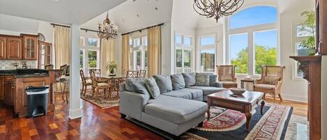 A spacious living room with a large sectional sofa, a dining area with chairs, a kitchen with wooden cabinets, and a large window offering a view of greenery.