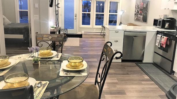 A modern kitchen with a dining area, featuring a glass-top table, chairs, and a view of the outside through large windows. - Esther's Place-Carleton Place Downtn Apt (Carleton Place)