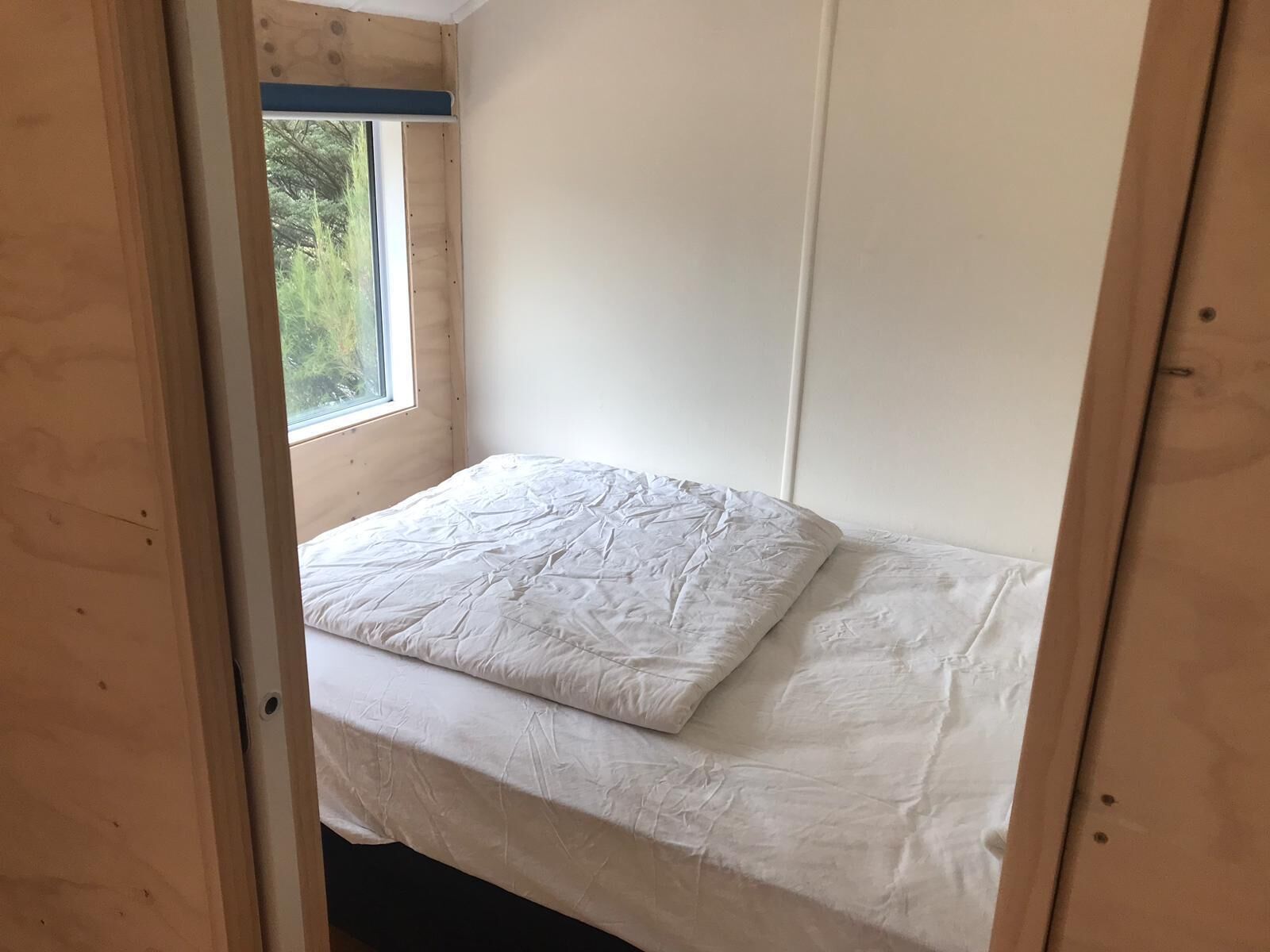 A small, single-bed room with a window, wooden walls, and a white mattress.