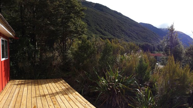 Une terrasse en bois, avec un bâtiment rouge, entourée d’une végétation luxuriante et avec des montagnes en arrière-plan.