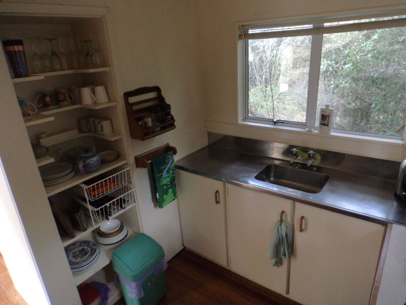 A compact kitchen with white cabinets, a stainless steel countertop, a window with a view of greenery, and a small trash bin.