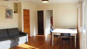 Un salon avec un canapé gris, un plancher en bois, une table blanche avec des chaises et une porte donnant sur une autre pièce.