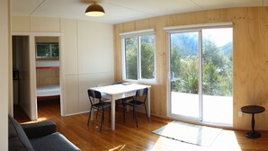 Une pièce au décor intérieur en bois, une table blanche avec des chaises, un banc et une grande porte-fenêtre coulissante donnant sur un balcon avec vue sur la verdure.