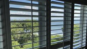 A window with horizontal blinds, offering a view of a green landscape and a body of water. - Bay Breezes 2BR / Pet-Friendly Cedars East RVA (Longboat Key)