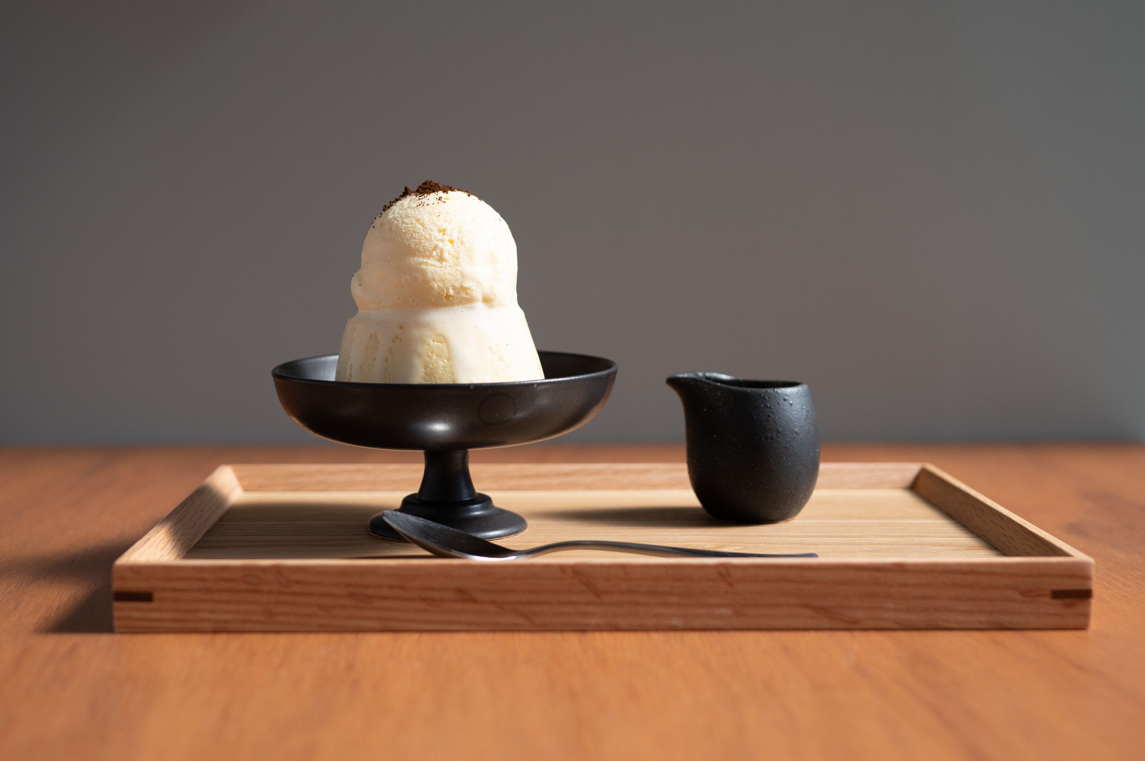 A dessert in a black bowl on a wooden tray with a spoon and a small black cup beside it.