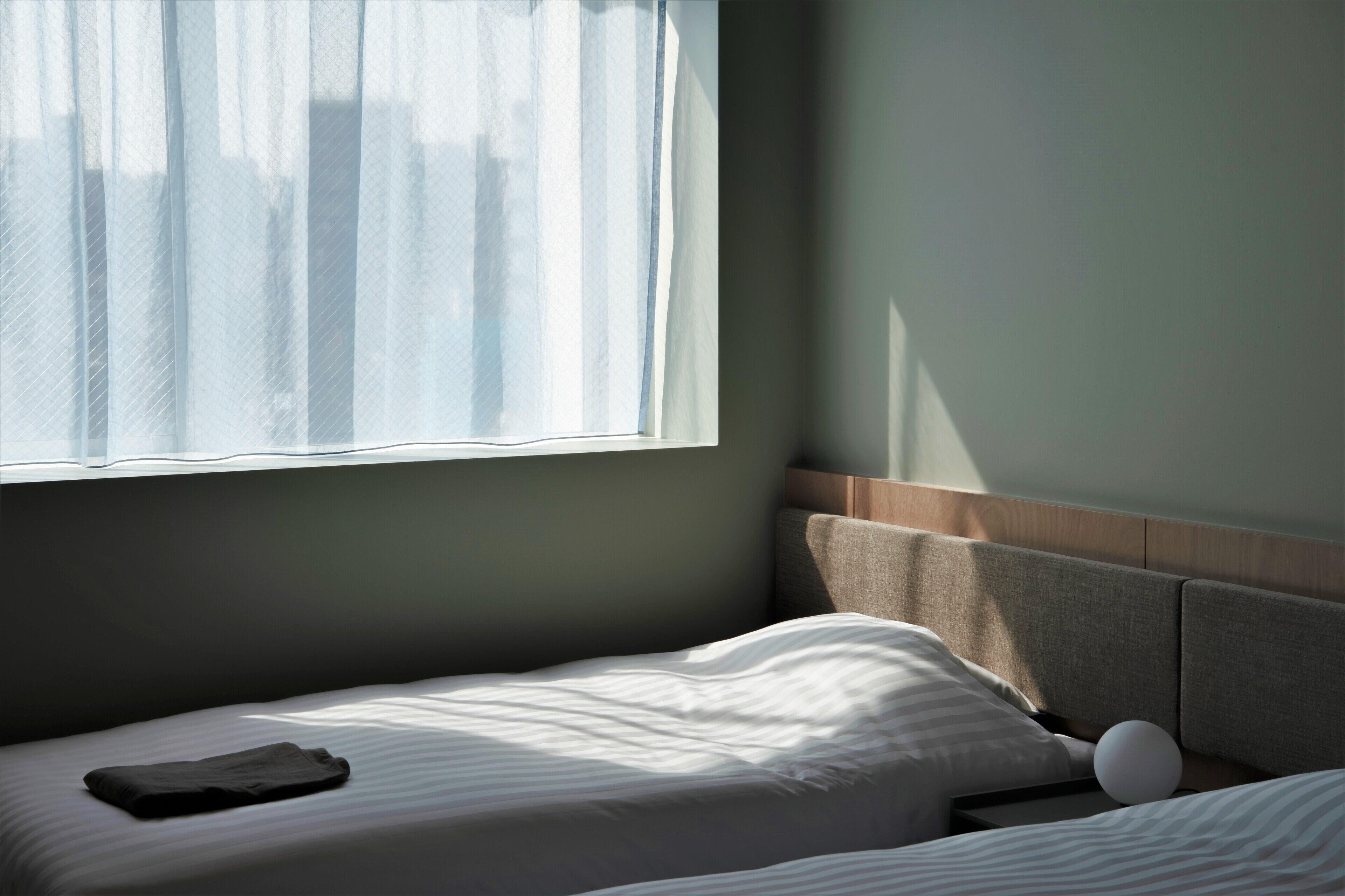 A neatly made bed with a pillow and a folded garment, a bedside table with a spherical object, and a window with sheer curtains allowing natural light.