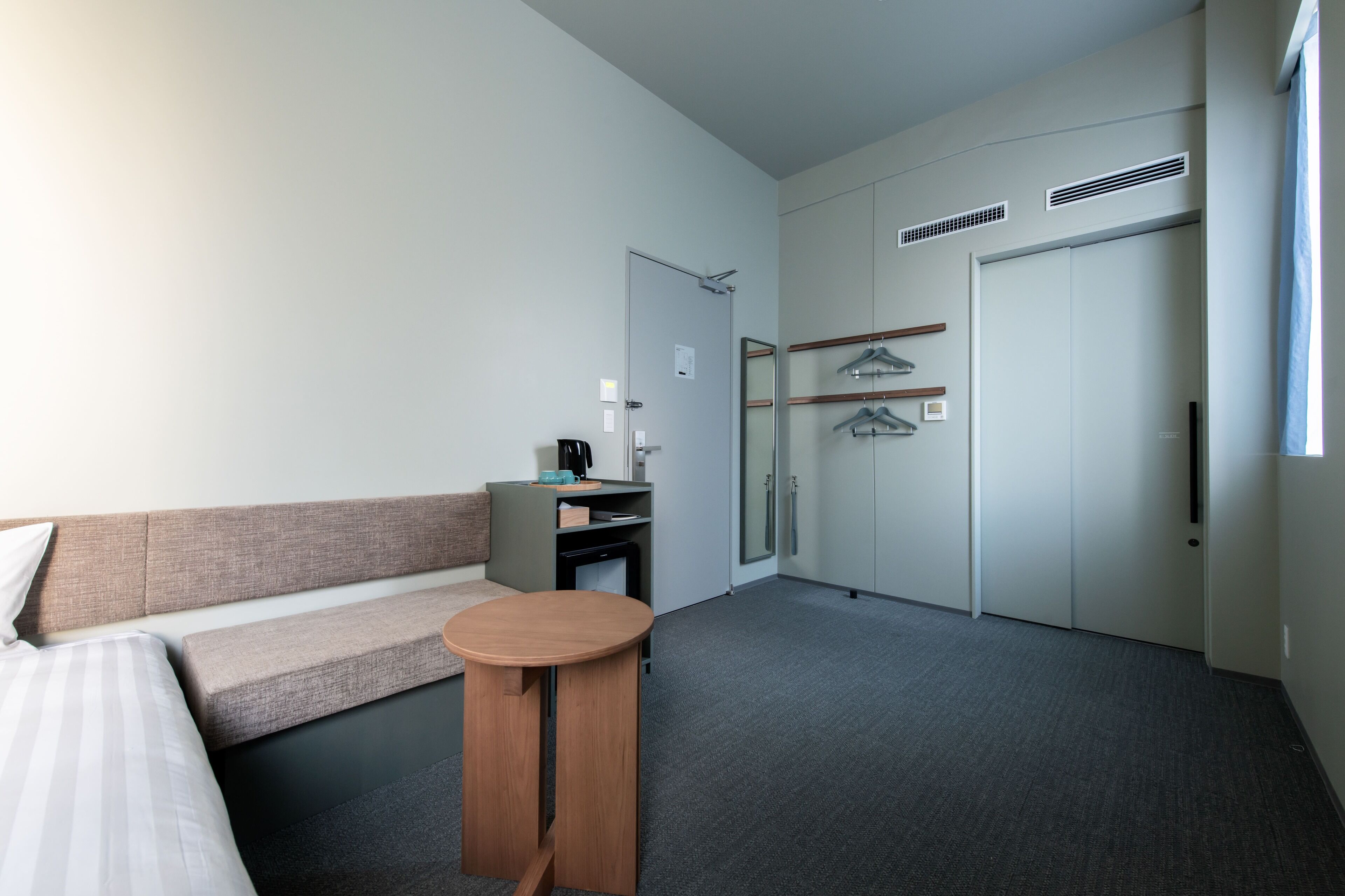 Une chambre d’hôtel comprenant un lit, une petite table, un canapé, un bureau avec une chaise, une armoire et un porte-manteau.