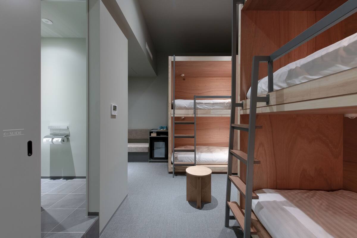 A modern hotel room with bunk beds, a small wooden stool, a desk, and a toilet.