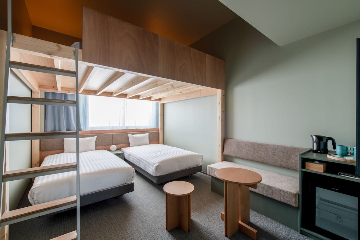 A compact hotel room with bunk beds, a small seating area, a kitchenette, and a window providing natural light.