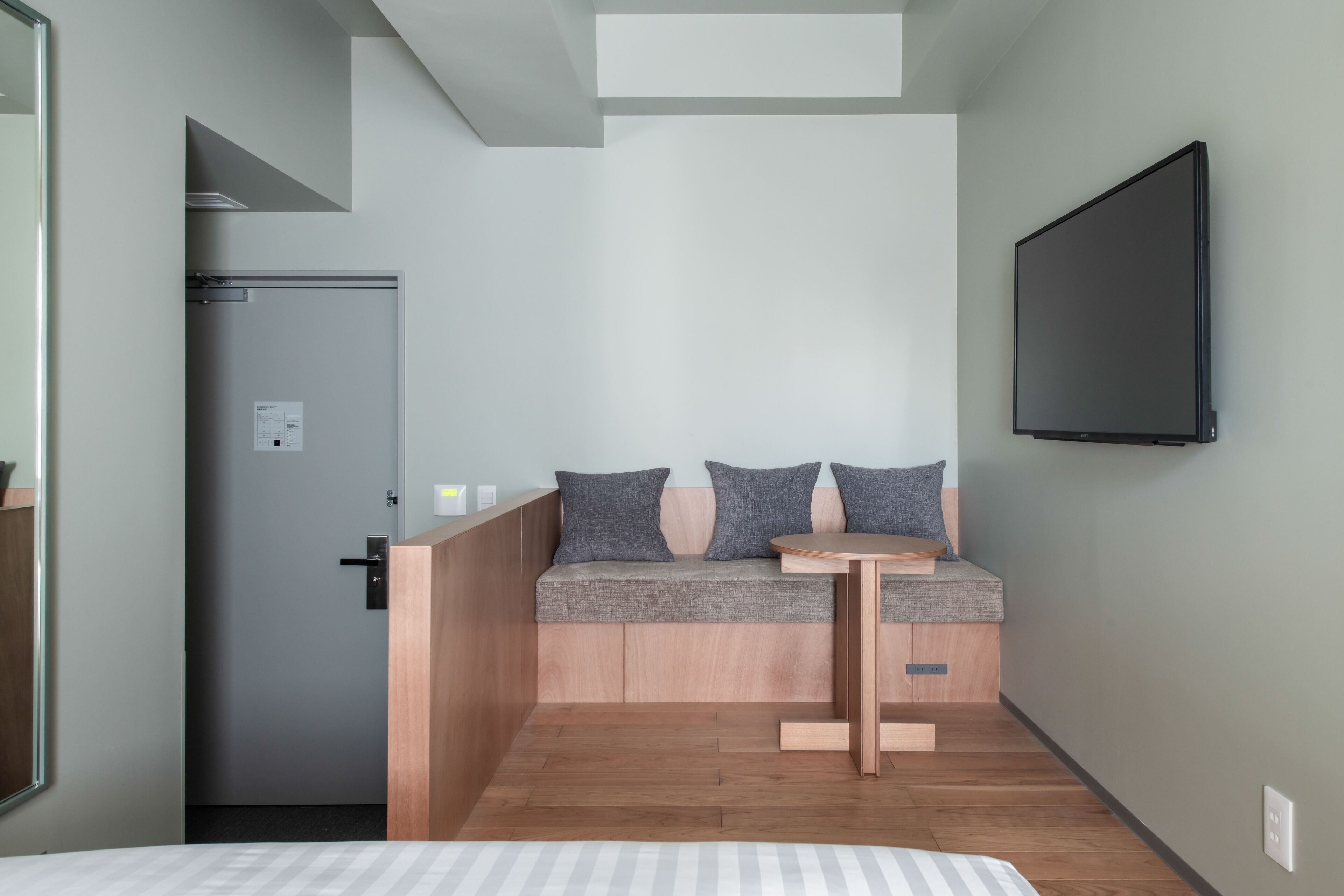 Habitación de hotel moderna con un banco de madera, una pequeña mesa redonda y un televisor de pantalla plana montado en la pared.