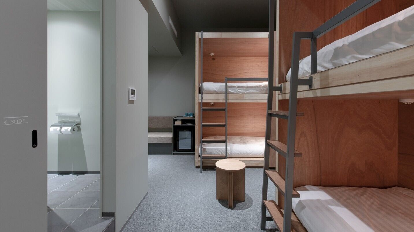 A modern hotel room with bunk beds, a small round table, and a visible bathroom with a toilet and towel dispenser.