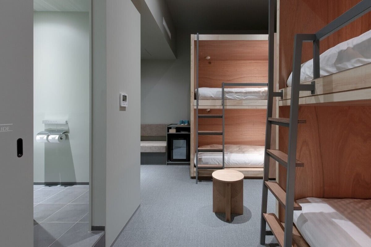 A modern hotel room with bunk beds, a small round table, and a visible bathroom with a toilet and towel dispenser.