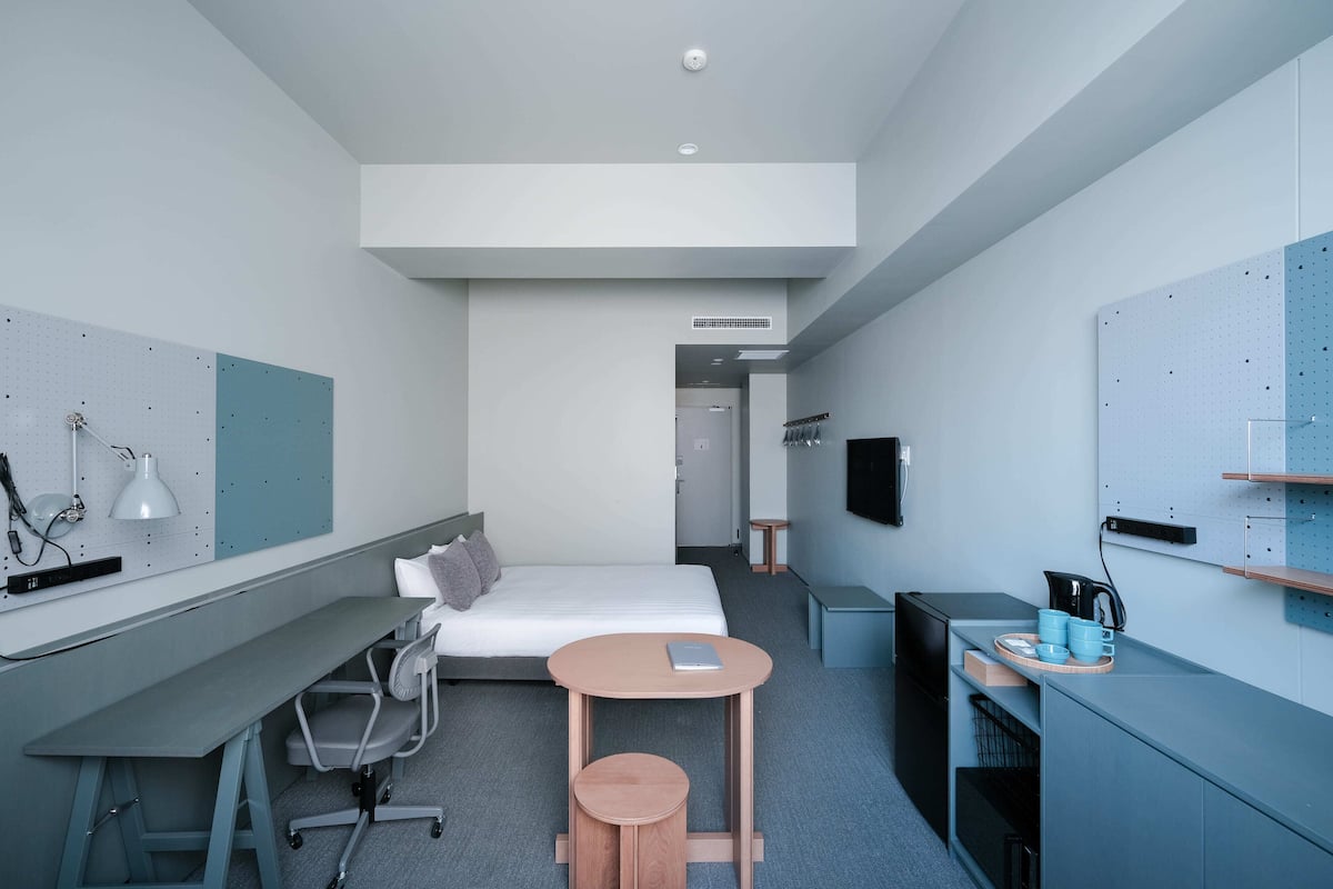 A compact hotel room with a bed, desk, chair, small round table, and a TV mounted on the wall.