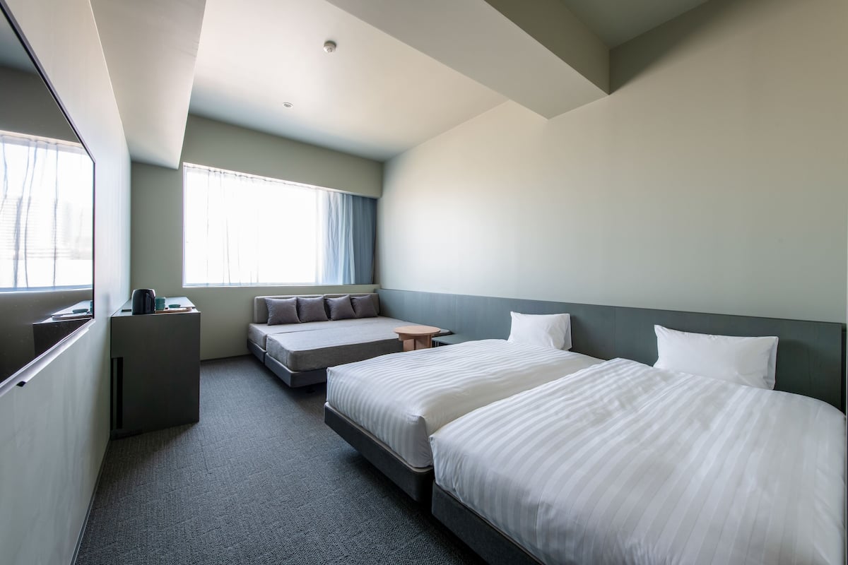 A modern hotel room with two beds, a sofa, a small table, and a window with curtains.